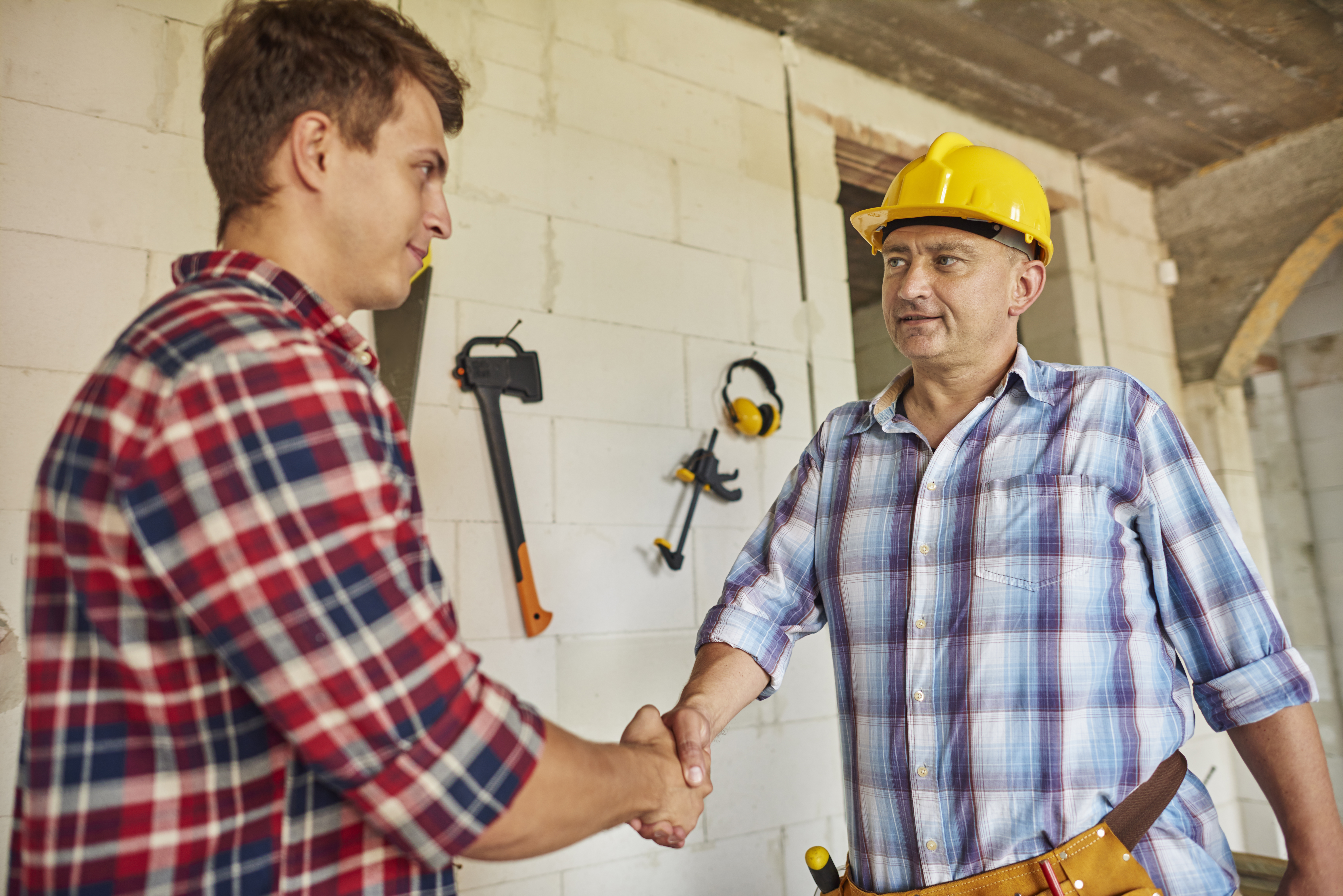 One of the Best Construction Contractors in Ottawa, wearing a tool belt, plaid shirt and yellow hardhat confidently shakes the hand of his male client, who is also wearing a plaid shirt.  Both are standing in an unfinished concrete block walled room.