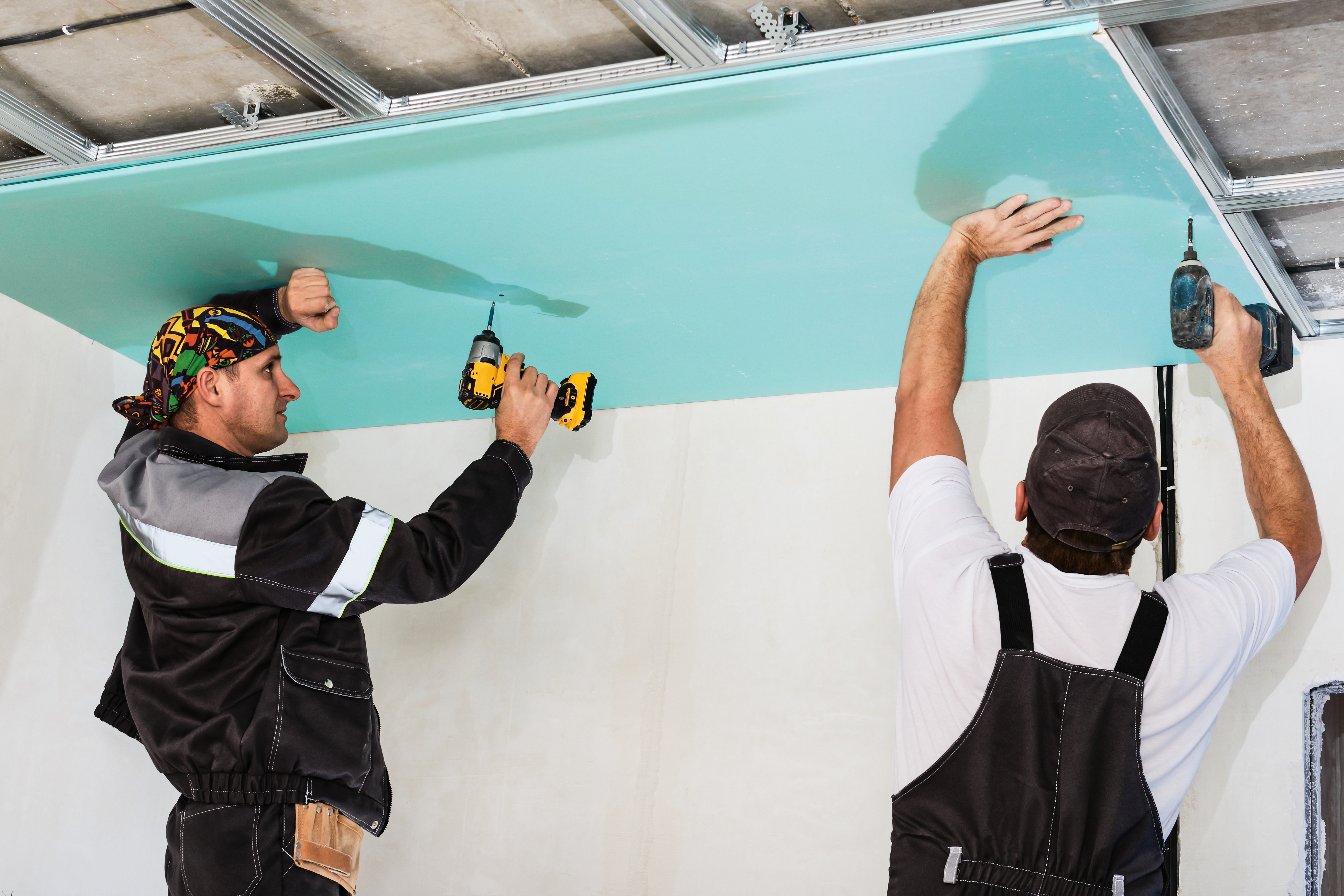 Two installers working for The Best Drywall Contractors In Ottawa. Each are wearing black work coveralls, and are holding a single sheet of drywall against a ceiling, and each are using cordless drills to secure the sheet in place with screws.