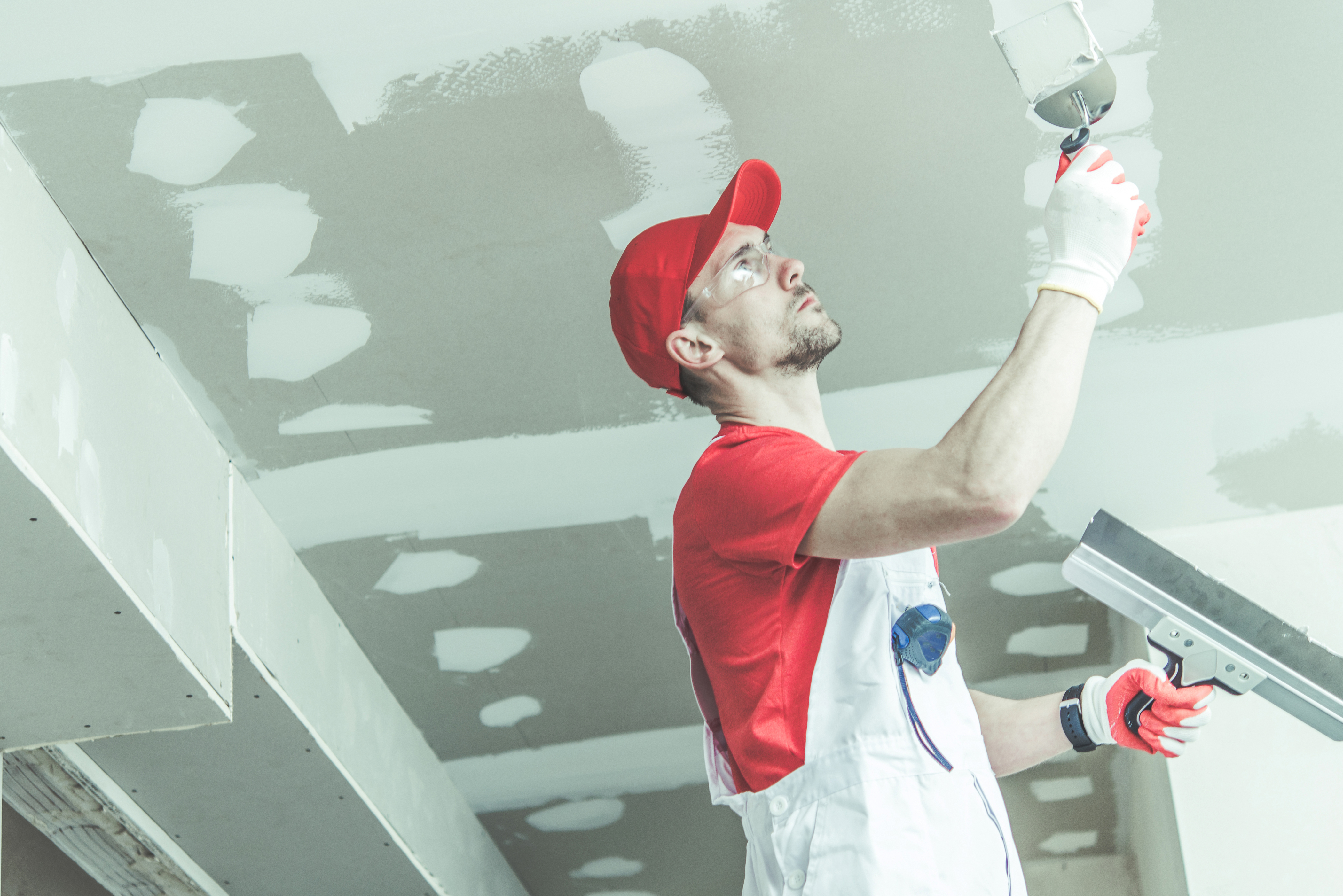 One of the members of the Best Drywall Finishers In Ottawa, wears a red cap, red shirt, and white coveralls, and is applying drywall compound to drywall / gyprock ceiling joints. In each hand, the finisher is holding different sized drywall trowels.