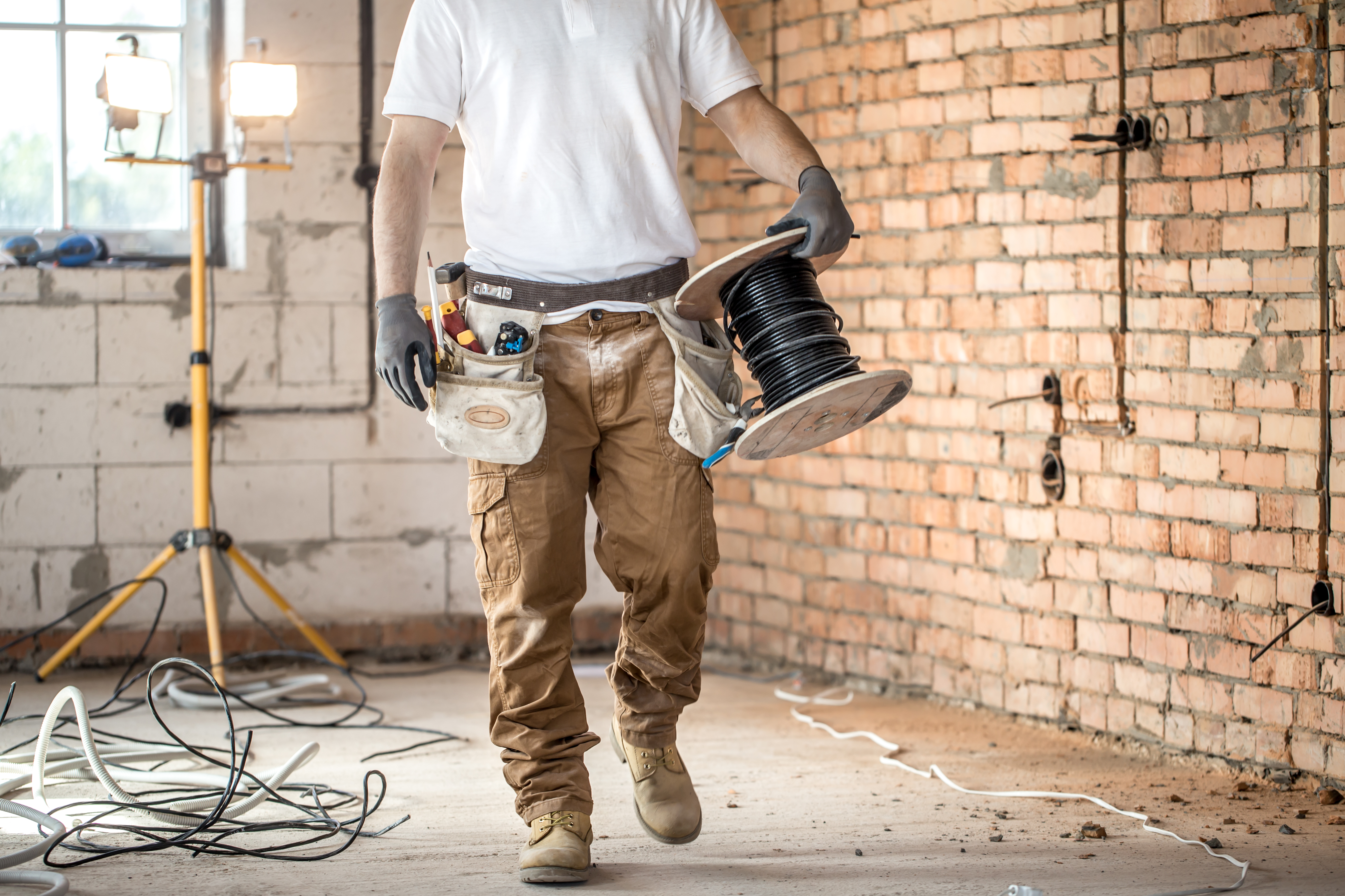 A Top-Rated Electrician in Hamilton, dressed in a white shirt, brown construction trousers, a full tool belt and work boots, can be seen carrying a wood spool of black wire across a brick and block room that is obviously part of a construction project.