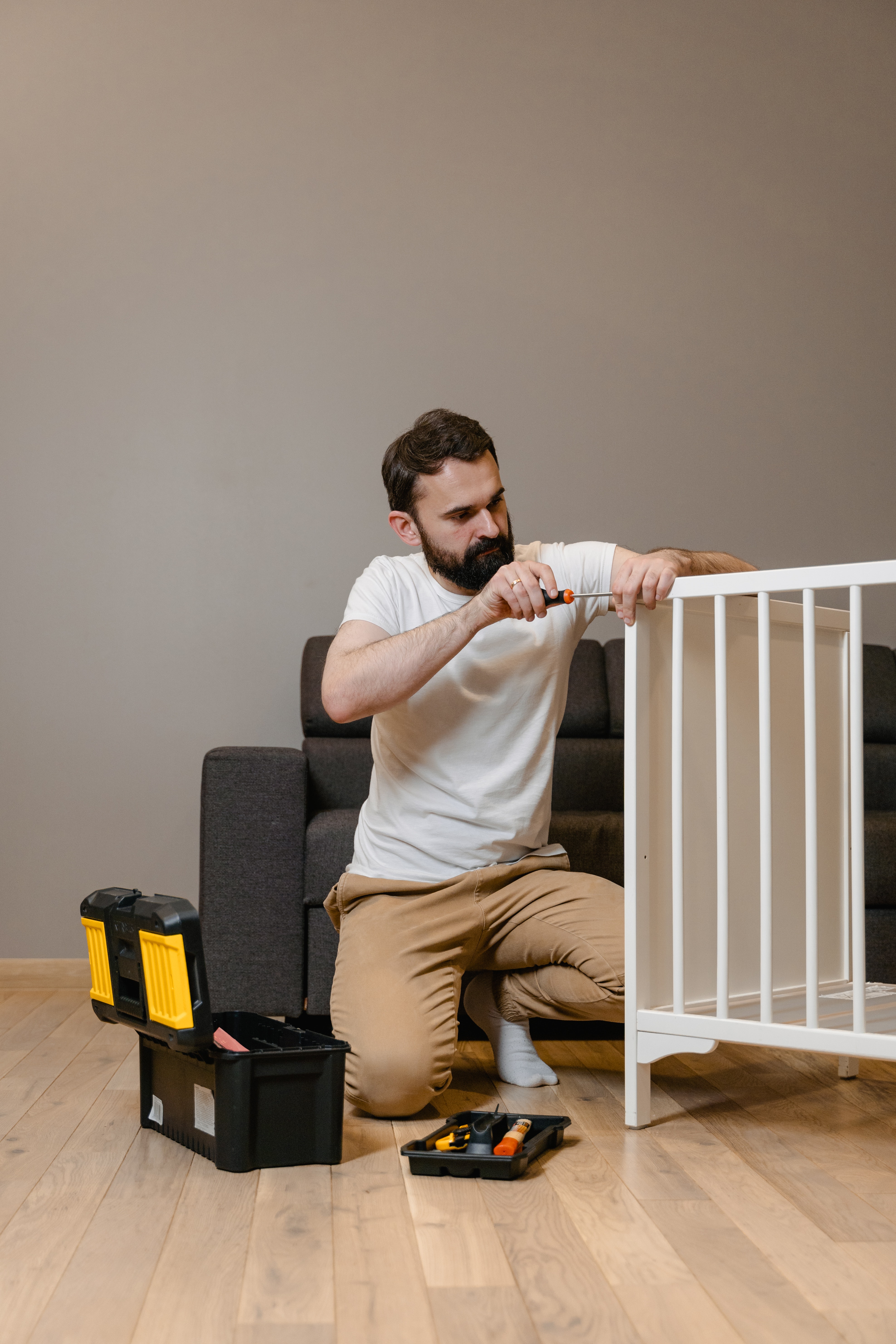 An employee from one of the Best Handyman Service Providers in the Hamilton Region, kneels beside his open toolbox while working to assemble a baby’s white coloured crib.