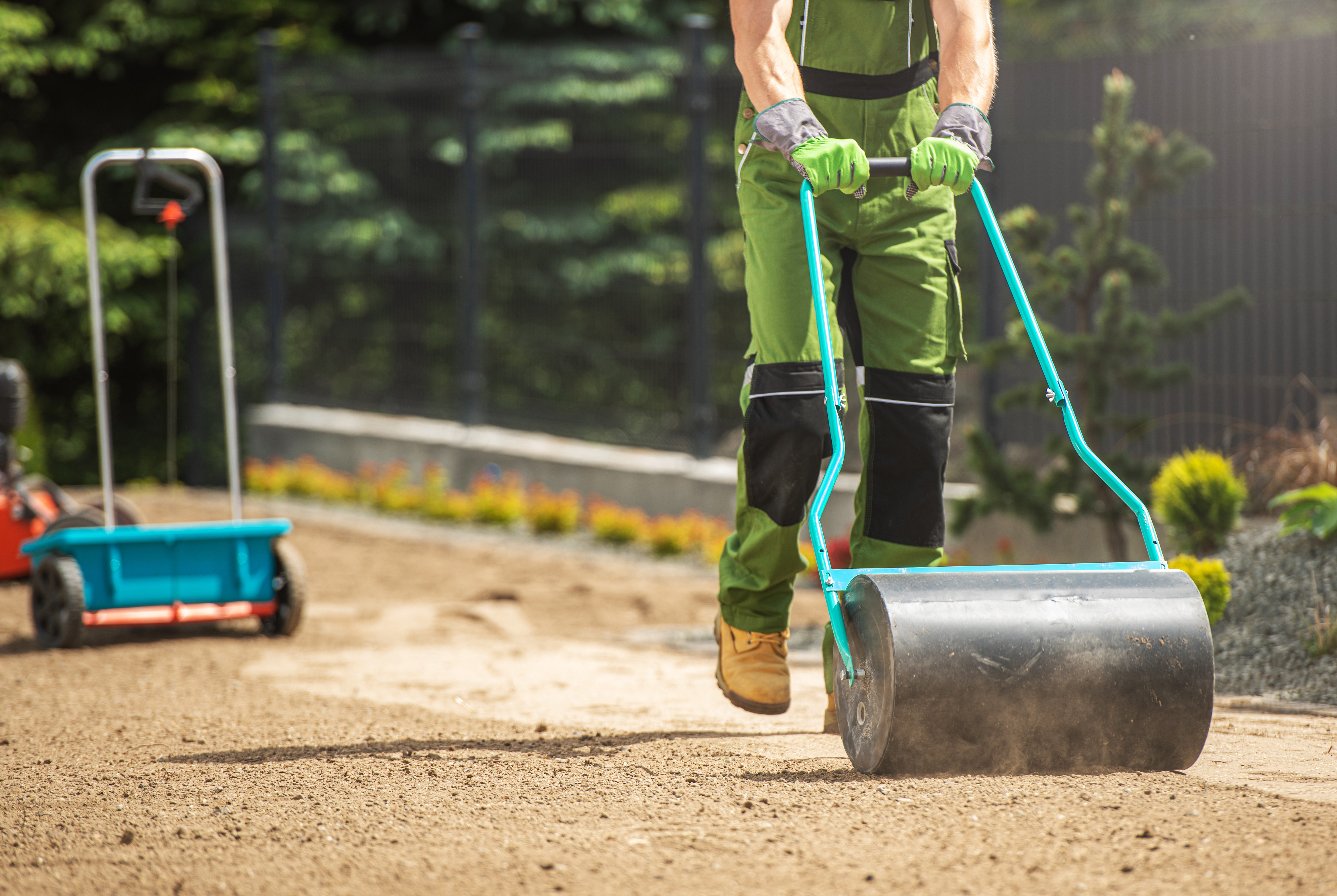 A crew member from one of the Best Landscaping Contractors in the Hamilton Area can be seen dressed in green work trousers with black knee pads. He is pushing a lawn roller over crushed gravel seen in a pathway setting. In the background, there is a green forest and a spare seed dispenser on wheels.