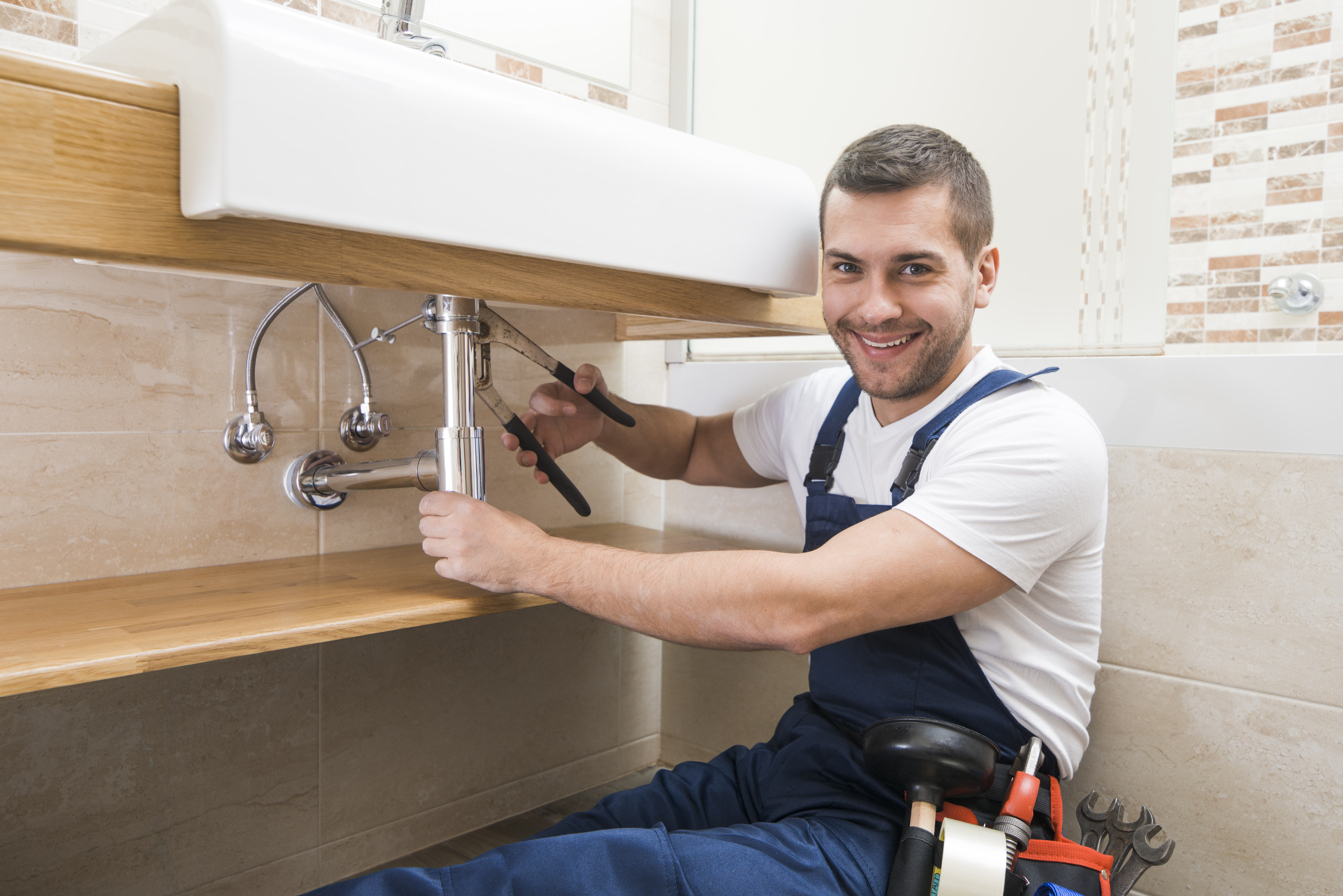 A Top Rated Plumbing Contractor in Ottawa, wearing a white shirt and blue coveralls is shown sitting parallel to a sink drain.  He is holding a valve stem in one hand and a set of pliers in the other while working on hooking up the drain.