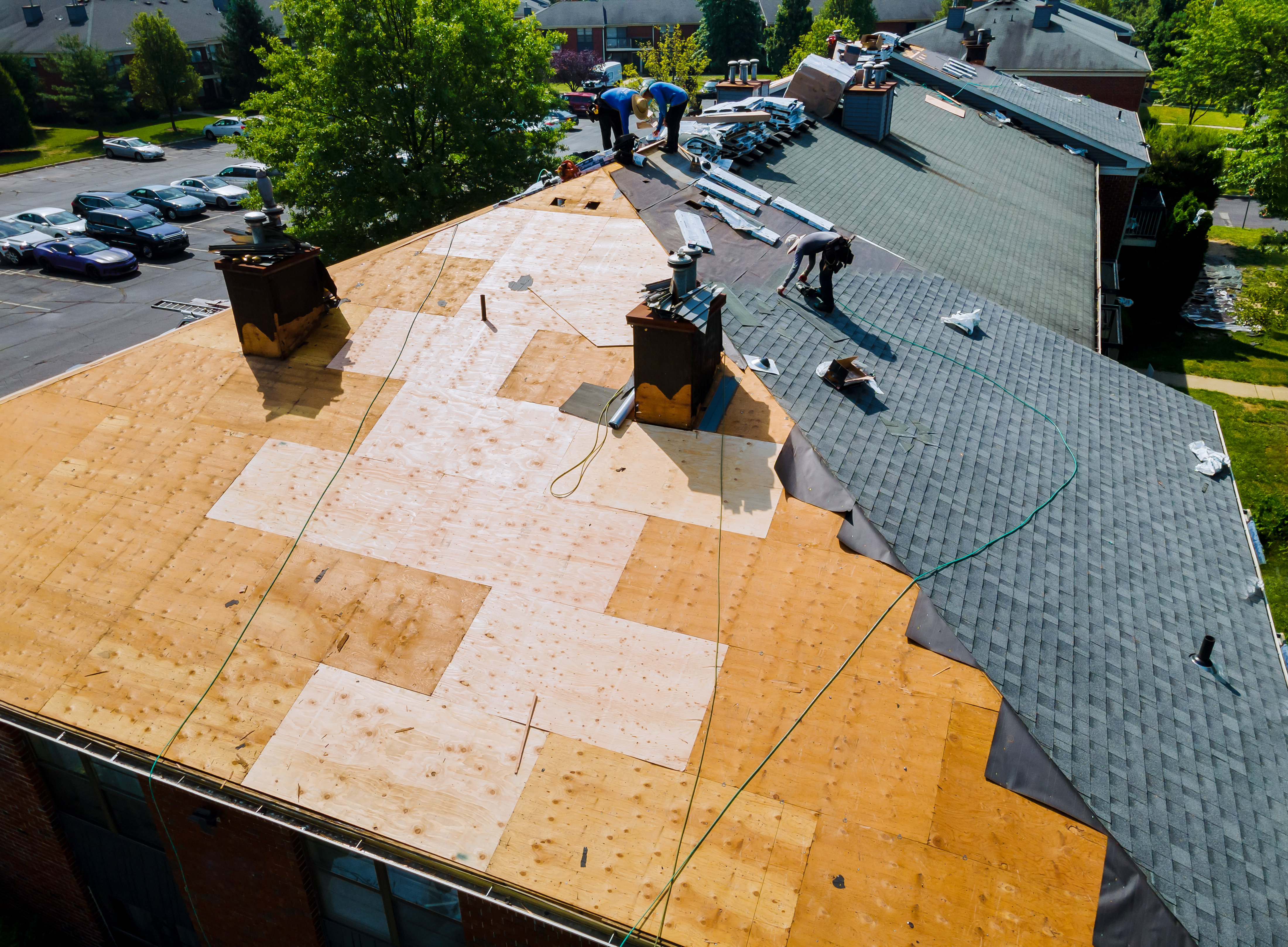 Professional Roofing Contractors in Hamilton are standing on a multi unit, multi pitched roof working to install dark shingles. Half the roof has been shingled; the other half shows the bare plywood sheets ready to be covered. Roll roofing products and packages of shingles appear in the background.